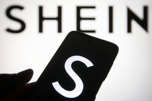 Na imagem, logo da Shein ao fundo com mãos segurando celular com S na tela - Metrópoles