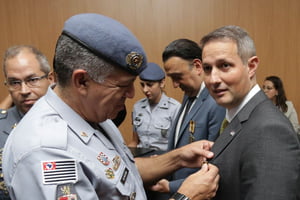 Imagem colorida mostra Guilherme Derrite, homem grisalho, branco, de terno cinza, recebendo uma medalha de um oficial fardado da PM - Metrópoles
