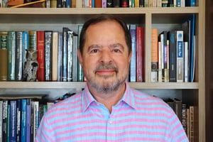 Imagem colorida mostra Sergio Machado Rezende, um homem branco, com barba e bigode grisalhos, vestindo uma camisa polo listrada em azul e rosa na frente de um estante cheia de livros - Metrópoles