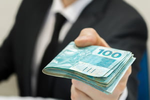 Foto colorida de empresário estindo terno preto e gravata segurando notas de dinheiro no valor de R$100 homens brancos - Metrópoles