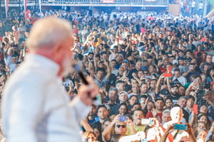 Imagem colorida mostra Lula, de costas, desfocado, com uma multidão de pessoas na sua frente