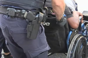 Imagem colorida de policial carregando um homem em uma cadeira de rodas preso por tráfico de drogas - Metrópoles