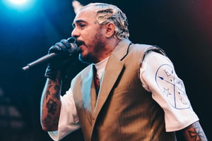 Homem pardo com cabelo descolorido e arrumado em ondas com gel segura microfone em palco, ele veste colete marrom sobre camiseta branca