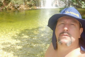 Foto colorida de homem branco, com sobrepeso e cavanhaque, usando chapéu em dia ensolarado em frente à cachoeira de águas esverdeadas - Metrópoles