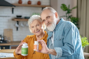 Foto colorida de mulher e homem idosos segurando uma embalagem de medicamento. Eles estão em uma cozinha - Metrópoles