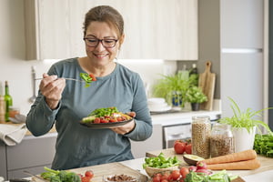 Foto colorida de mulher comendo legumes. Ela está perto de uma mesa com legumes e verduras - Metrópoles