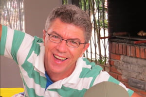 Homem branco, sem barba, sorri com boca aberta e usando óculos em sala de casa - Metrópoles