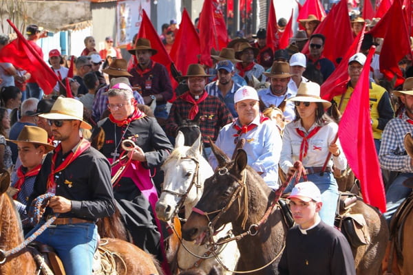 Foto colorida tirada de dia de pessoas montadas à cavalo em meio à multidão carregando bandeiras vermelhas