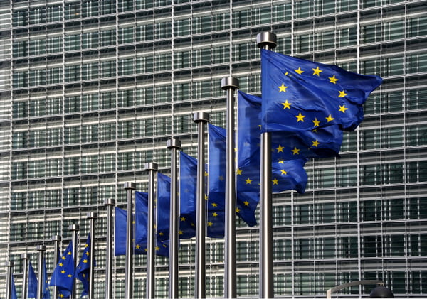Imagem colorida de bandeiras azuis com estrelas amarelas da União Europeia, dispostas uma ao lado da outra, tremulando, com um prédio ao fundo - Metrópoles