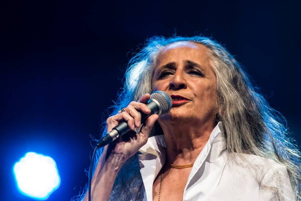 Foto colorida da cantora Maria Bethânia. Ela segura um microfone com a mão direita e usa uma blusa branca. Tem cabelos grisalhos - Metrópoles