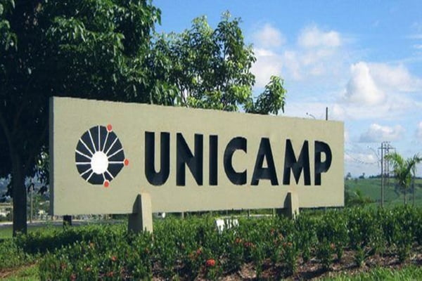 Imagem colorida mostra placa da Unicamp. O objeto é feito de concreto é mostra a logo da universidade seguida pela sigla. Ela está cercada de plantas | Metrópoles