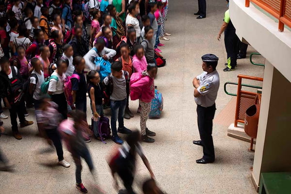 imagem colorida mostra adolescentes em escola usando uniforme em frente a militar da PM, que usa farda
