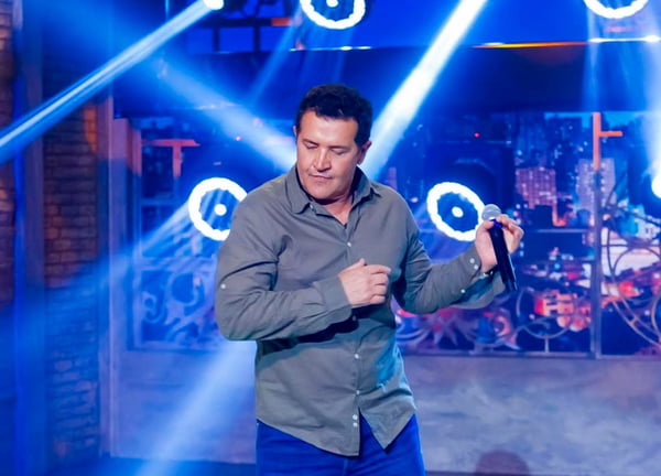 Foto colorida do cantor Beto Barbosa. Ele est&aacute; no palco, com um microfone na m&atilde;o, e usa camisa de bot&atilde;o verde escuro - Metr&oacute;poles