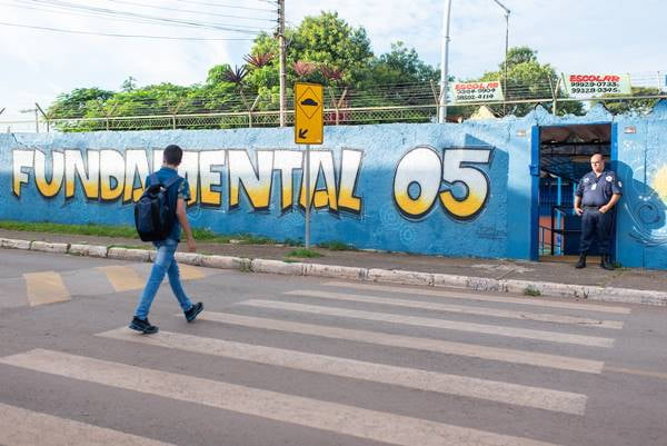 Menino com mochila nas costas atravessando rua em faixa de pedestre e muro azul de escola do outro lado