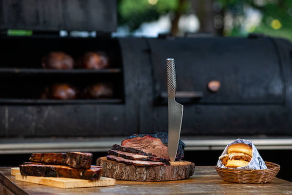 Cortes de carne e hambúrguer sobre mesa de madeira com defumadora ao fundo
