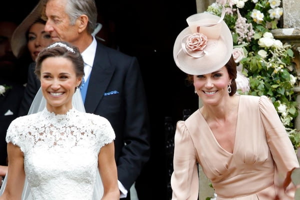 Foto colorida de mulher com vestido de noiva ao lado de outra, com chapéu e vestido no tom rosé. As duas estão sorrindo - Metrópoles