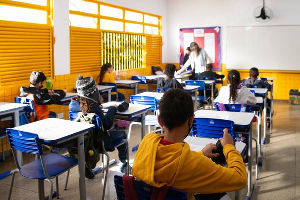 Sala de aula com alunos de costas, sentados em carteira, olhando para professora à frente da sala; janelas amarelas