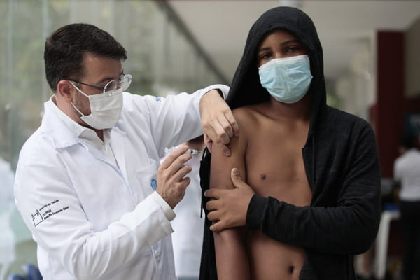 Secretário de Saúde do Rio, Daniel Soranz, aplica vacina em um adolescente. O médico é um homem branco, de jaleco branco, com óculos e máscara branca. O jovem é negro, com um casaco preto e de máscara azul