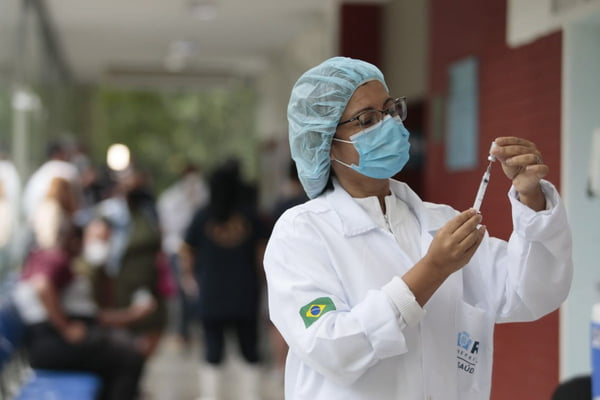 Vacinação na cidade do Rio de Janeiro segue intensa para imunizar toda população contra Covid-19. Na foto, uma enfermeira branca, com jaleco e touca branca, óculos de grau e máscara azul segurando uma seringa de vacina