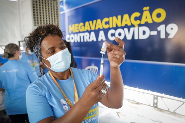 Mulher negra com seringa na mão e banner azul de vacinação contra a Covid-19 atrás