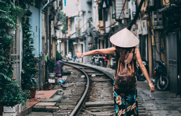Na foto, uma mulher pedindo carona com chapéu chinês no meio de uma cidade, em cima do trilho do trem