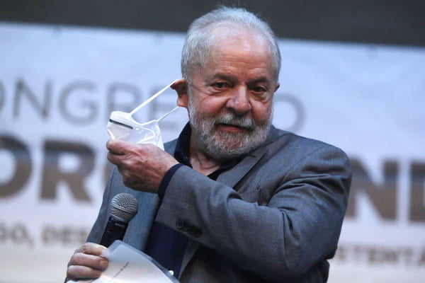 Lula tira a máscara para falar em evento. Ele segura um microfone e usa terno - Metrópoles