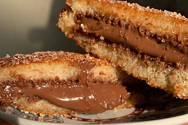 Na foto vemos um sanduíche de pao branco com recheio marrom escuro que se assemelha a nutella e por cima dos dois alguns cristais de açúcar