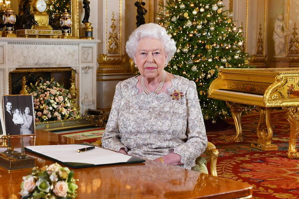 Mulher com cabelos brancos sentada. Ao fundo, uma árvore de Natal e um piano. Na mesa, há um porta-retrato