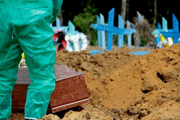 Fotografia colorida. Homem enterra caixão marrom em cemitério. Ao fundo, cruzes pintadas de azul sinalizam outros caixões enterrados