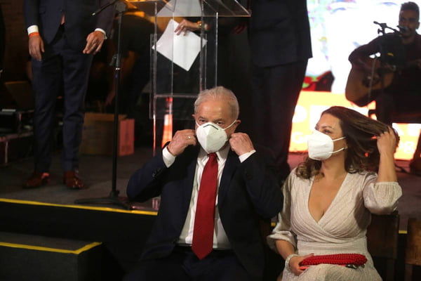 Em evento, Lula coloca a máscara, sentado próximo ao palco do lado de sua esposa, Rosângela. Ela também usa máscara e arruma o cabelo - Metrópoles