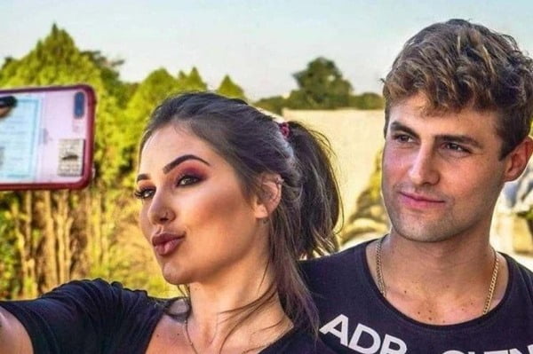Na imagem colorida, duas pessoas estão posicionadas do centro para a direita. Eles usam roupas escuras e posam para fotografia feita por um celular