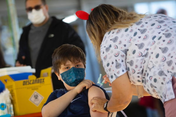 Menino com máscara azul, puxando a manga da camisa e mulher de costas aplicando vacina no braço dele