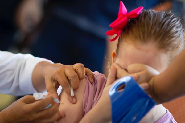 Menina com laço rosa no cabelo com a mão no rosto e mão de enfermeira aplicando vacina no braço dela