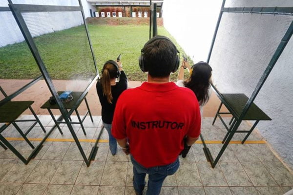 Homem de costas com camisa vermelha escrito "Instrutor" e duas mulheres de preto, de costas, apontando armas para a frente
