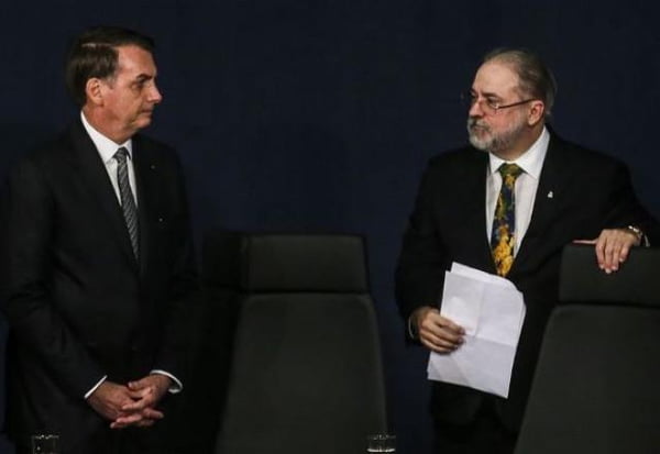 O presidente Bolsonaro e o procurador Augusto Aras em evento. Eles estão em pé, próximos às cadeiras, e se encaram. Ambos usam terno, mas estão sem máscara - Metrópoles