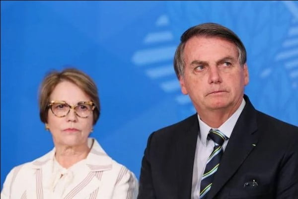 Bolsonaro e Tereza Cristina, ministra da Agricultura, sentam-se juntos em evento no Planalto. Ela usa óculos, tem cabelos curtos e loiros. Ele usa terno e gravata, camiseta clara. Ambos olham seriamente para frente - Metrópoles