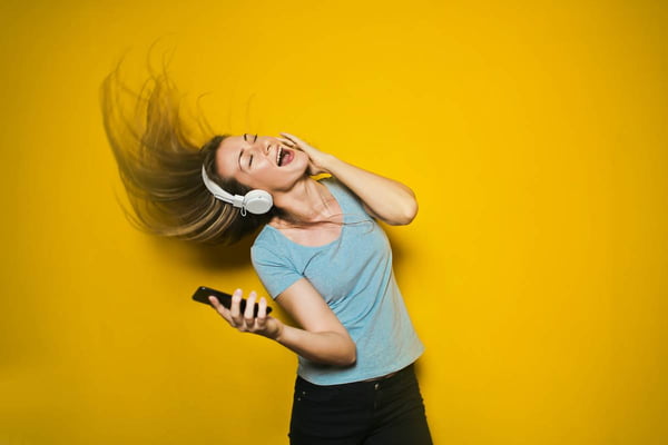 Uma mulher aparece na frente de um fundo amarelo com camiseta cinza de fones de ouvido branco cantando e dançando