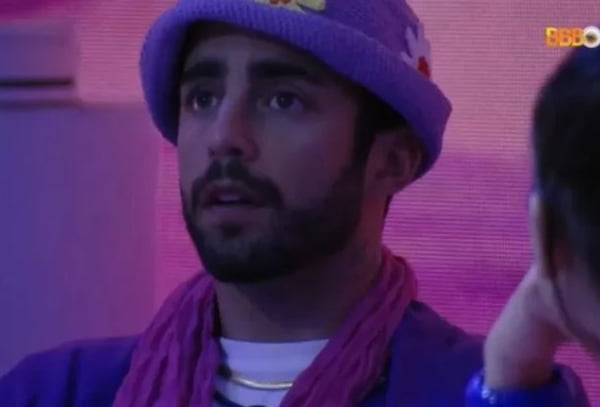 Pedro Scooby tem pele clara, barba escura e usa roupa e chapéu lilás