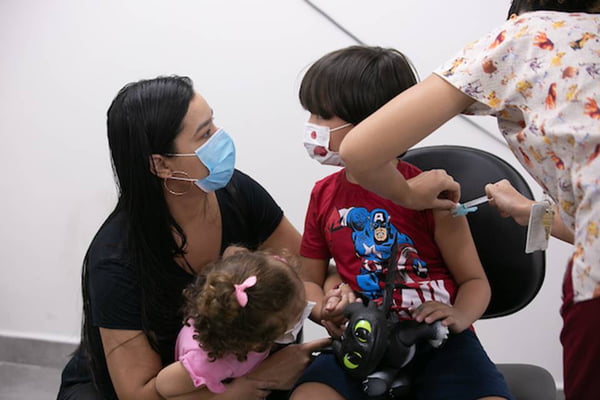 imagem colorida mostra criança sendo vacinada usando mascara de pano e sentada numa cadeira, ao lado da mãe também de mascara e de uma criança menor - Metrópoles