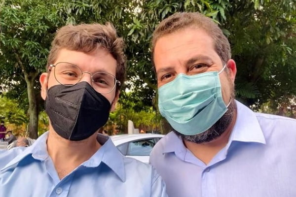 Dois homens de camisa social azul e máscaras sorrindo para foto