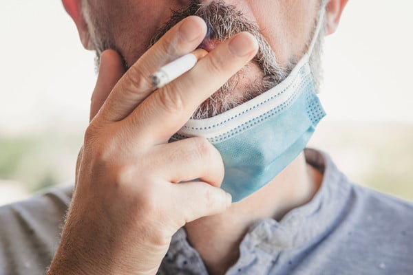 Homem fuma cigarro e segura com a mão. Ele tem uma máscara no queixo