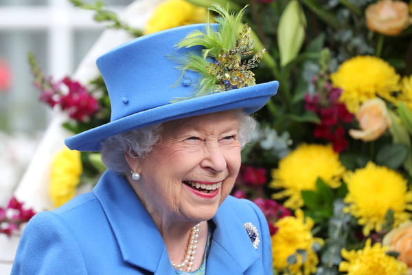 Foto colorida. Rainha Elizabeth com roupas azuis. Ao fundo da imagem, tem flores
