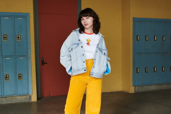 Mulher branca com calça amarela, camiseta branca e jaqueta jeans. Ela está no corredor de uma escola. Ao fundo é possível ver a porta de uma sala de aula e armários. As roupas são da marca Levi's com o desenho Os Simpsons.