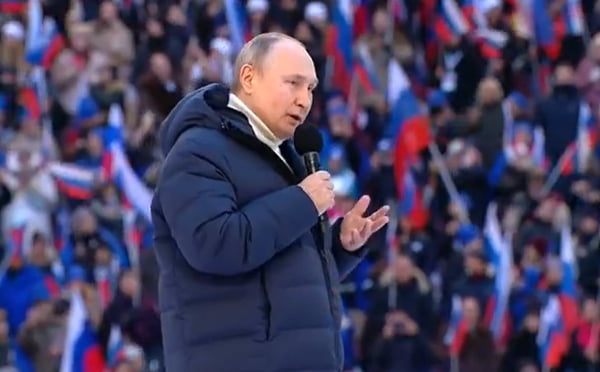 Presidente da Rússia, Vladimir Putin cita versículo bíblico ao defender invasão da Ucrânia em discurso num estádio. Ele usa casaco de frio e segura microfone, de perfil - Metrópoles