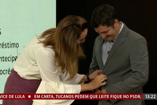 A apresentadora da Globo News Maria Beltrão ajuda convidado que deixou cair o microfone. Na foto, ela ajeita o microfone no terno dele - Metrópoles