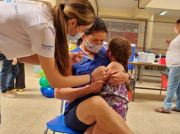 Criança sentada em colo de mulher, com a cabeça para trás enquanto mulher aplica vacina em seu braço