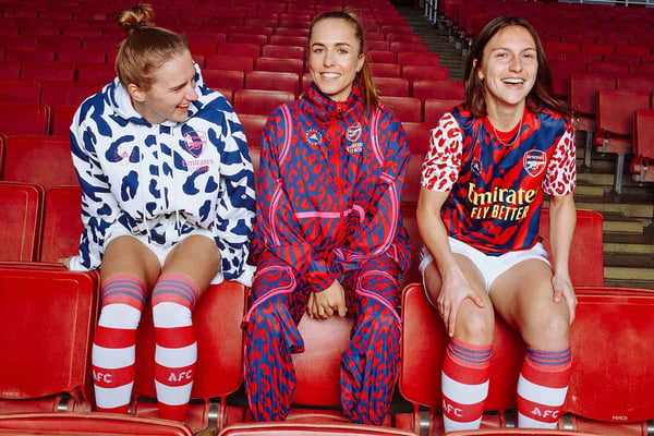 Jogadoras do time feminino de futebol inglês Arsenal sentadas em bancos vermelhos da arquibancada de um estádio. Ela usam roupas da marca Adidas em colaboração com a estilista Stella McCartney.