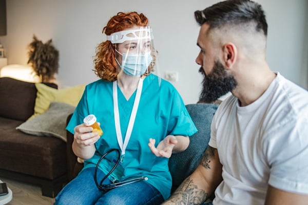 Mulher vestida com roupa que remete a profissionais da área da saúde, segurando um recipiente laranjado de medicamento e utilizando máscara contra a Covid ao lado de um homem com cabelo e barba pretos e camiseta branca- Metrópoles