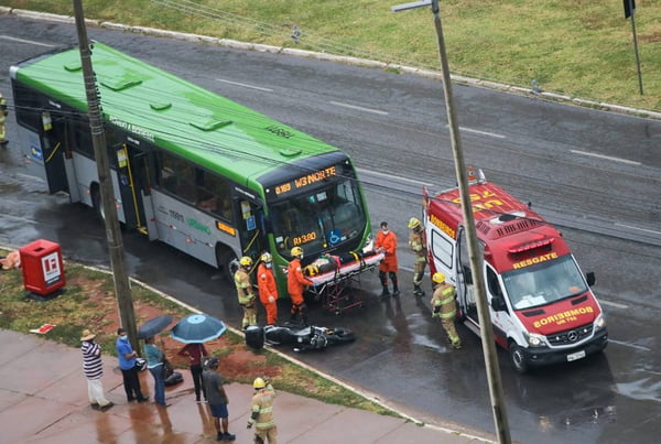 Ônibus verde parado diante de motocicleta caída ao cahão, com bombeiros prestando atendimento a motociclista ferido