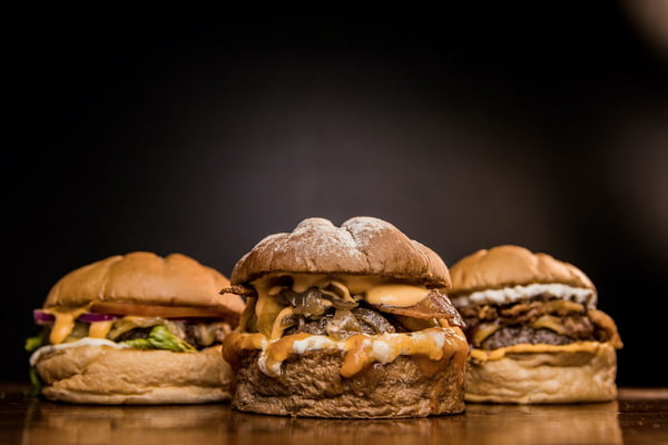 Foto com três hambúrgueres de carne e ingredientes variados, estando dois deles em segundo plano sob fundo preto - Metrópoles